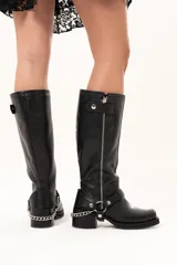 Bota estilo biker de cuero negro con caña alta y cierre metálico lateral. Presenta un diseño versátil con estribo y cadena desmontables, taco cuadrado y suela con plataforma.