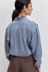 Camisa de satén color azul grisáceo, con cuello mao con detalle de volados, cierre frontal con botones y mangas largas con puños abotonados.