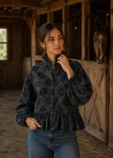 Camisa de lino con estampado floral en tonos azul y oliva. Presenta cuello alto con volados, cierre frontal con botones forrados, mangas largas abullonadas con puños fruncidos y un detalle de volado en el ruedo.