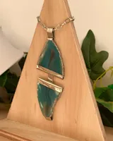 Collar con dije colgante de dos piezas de ágata teñida de color verde azulado, unidas por un conector metálico plateado. El dije tiene bordes irregulares y está engastado en metal plateado. La cadena es de eslabones gruesos plateados.