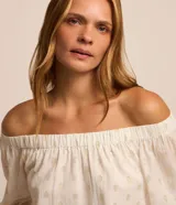 Blusa de tricolina blanca con estampado de lunares dorados en foil, escote descubierto estilo hombro a hombro y mangas largas abullonadas con puños elásticos.