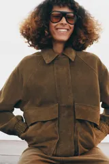 Chaqueta oversize confeccionada en piel de ante, con cuello solapa, manga larga con puños elásticos y bajo elástico. Presenta bolsillos de plastrón con solapa en el frente y cierre frontal con cremallera y botones ocultos.