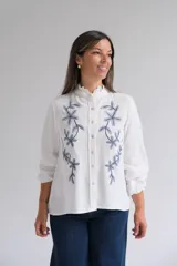 Blusa blanca de corte recto con cuello mao y volado pequeño, cierre frontal con botones y mangas largas con puños elásticos. Presenta un delicado bordado floral en color azul en la parte frontal.