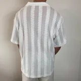 Camisa de tejido calado con textura, color blanco, de corte oversize, manga corta y cuello solapa con cierre frontal de botones.