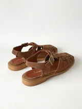 Sandalias estilo cangrejera color marrón, confeccionadas en cuero con diseño trenzado en la parte delantera, tira ajustable con hebilla metálica en el tobillo y suela plana.