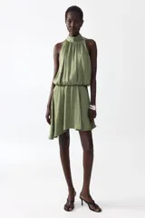 Vestido corto verde oliva con leve brillo, cuello halter elevado con frunces, cuerpo holgado con cintura elástica y falda corta de corte asimétrico.
