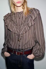 Camisa de manga larga con cuello subido, cierre frontal con botones y detalle de volantes en el pecho. Presenta un diseño a rayas verticales y puños elásticos.