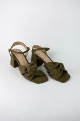 Sandalia de taco cuadrado medio, confeccionada en material textil símil gamuza color verde oliva. Presenta un diseño de punta cuadrada con una tira frontal anudada y una tira ajustable con hebilla metálica en el tobillo.
