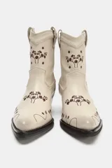 Botín estilo cowboy confeccionado en piel color crudo, con diseño de flores bordadas en tono marrón. Presenta tacón ancho en bloque, acabado en punta y tiradores laterales.