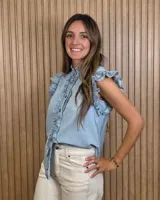 Musculosa de jean celeste sin mangas, con cuello alto fruncido, cierre frontal con botones y detalles de volados en las sisas y a lo largo de la botonera.