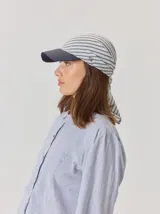 Gorro tipo pañuelo con visera rígida azul marino, confeccionado en lino con estampado de rayas grises y blancas. Presenta un diseño envolvente con caída trasera y detalle de logo bordado en el lateral.