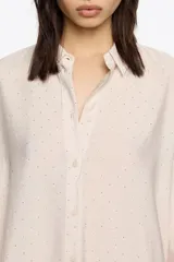 Camisa de corte recto en color beige claro, confeccionada en tejido fantasía con minibrillantes distribuidos por toda la prenda. Presenta cuello camisero y mangas largas con puño abotonado.