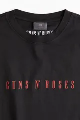 Remera negra de punto de algodón de corte holgado con hombros caídos, cuello redondo acanalado y estampa frontal con el nombre de la banda Guns N' Roses en color rojo.