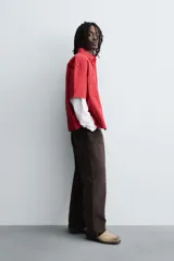 Camisa de algodón con estructura canvas, color rojo lavado, de corte relajado (relaxed fit) y manga corta. Posee cuello solapa, dos bolsillos de plastrón en el pecho y cierre frontal de botonadura. Presenta bordado y estampado de texto a contraste en el delantero.