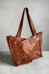 Cartera tipo tote de cuero natural color suela, con diseño envolvente, asas largas de hombro, bolsillo exterior y llavero metálico decorativo con logo.