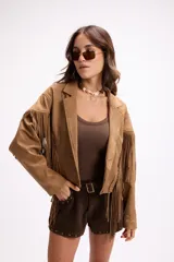 Chaqueta de efecto gamuza en color tostado, con solapas amplias, cierre frontal con un botón y flecos largos decorativos en la parte frontal y trasera de las mangas. Presenta una estructura definida y forro interior.