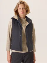 Chaleco gris con cuello alto y capucha, cierre frontal con botones a presión. La parte delantera es de lana gris lisa y la parte interior del cuello y el forro son de tela con patrón de espiga en blanco y negro.