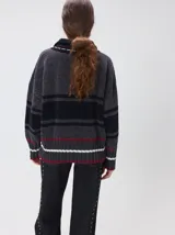 Pullover de punto con textura aterciopelada, diseño oversize, cuello polo con cierre frontal parcial y detalles de pespuntes en contraste. Presenta rayas horizontales en tonos tierra y un parche bordado con la letra M en el pecho.