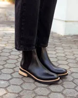 Botinetas tipo Chelsea de cuero negro, con paneles laterales elásticos y detalle de textura grabada en el talón. Presentan una suela plana con plataforma de madera clara y base de goma negra.