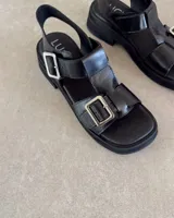 Sandalias de cuero color marrón con suela de goma negra, tiras anchas y hebilla metálica.