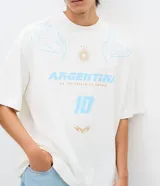 Remera de corte oversized color blanco, con cuello redondo y manga corta. Presenta una estampa frontal temática de Argentina en tonos celestes y dorados, que incluye el número 10, un sol y detalles gráficos tribales.