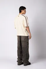 Remera de tejido de punto con textura calada, color crema con rayas verticales sutiles. Presenta cuello en V, mangas cortas con franjas laterales en color marrón oscuro y logo bordado en el centro del pecho.