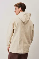 Campera canguro de gabardina color beige, con capucha ajustable con cordones, cierre frontal medio y bolsillo tipo ojal con solapa en el frente. Presenta mangas largas con puños elásticos y diseño holgado.