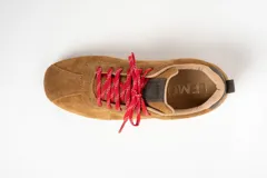 Zapatilla de estilo deportivo color camel, confeccionada en cuero gamuzado. Presenta cordones rojos con detalles blancos y un pequeño refuerzo gris en el talón.