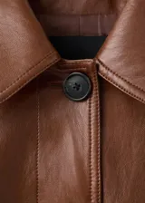 Chaqueta larga de efecto cuero color marrón, con cuello camisero, botonadura frontal oculta, bolsillos laterales y cinturón ajustable con hebilla en la cintura. Cuenta con trabillas en las muñecas y diseño amplio.