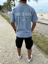 Remera oversize de color celeste melange, con diseño de estampado tipográfico en la espalda que incluye la frase "Time is Now" y otros detalles de colección.