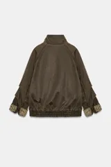 Chaqueta encerada color marrón con efecto lavado, cuello subido y manga larga. Cuenta con bolsillos frontales de plastrón y cierre de cremallera oculto por solapa con botones a presión. Los puños reversibles dejan ver un forro interior con estampado a cuadros.