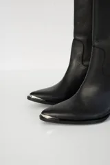 Bota bucanera de cuero color marrón oscuro, con taco cuadrado de 6 cm y punta fina con detalle metálico. Presenta una tira vertical decorativa con tachas metálicas en la parte superior de la caña.