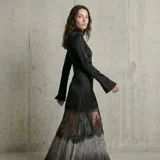 Vestido largo de satén negro con cuello alto y mangas largas. Presenta un diseño con una franja de encaje negro transparente en la parte inferior, que conecta el cuerpo de satén con una falda de satén en degradado gris oscuro.