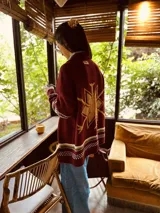 Cárdigan tejido de lana color borgoña con diseño de copo de nieve dorado en la espalda y bordados a mano. Presenta puños y ruedo con diseño geométrico en blanco, verde y dorado.