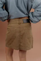 Minifalda de gabardina color tostado, de tiro medio con bolsillos delanteros y traseros, presillas para cinto y cierre frontal con botón metálico.