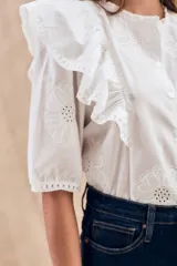 Blusa blanca de algodón con bordado floral, cuello redondo, manga corta con volados y cierre frontal con botones.