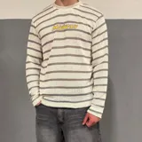 Sweater tejido de punto con diseño de rayas horizontales en tonos crema y gris. Presenta cuello redondo y el bordado de la palabra 'Midway' en color amarillo en el pecho.