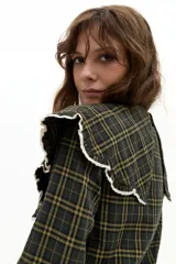 Camisa de manga larga confeccionada en popelina de algodón con estampado de cuadros en tonos verde oscuro y amarillo. Presenta un cuello amplio con bordes ondulados y terminaciones en encaje de algodón crudo, cierre frontal con botones de nácar y lazo de ajuste en el cuello.