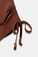 Chaqueta estilo safari de color marrón, con escote en pico, manga corta con cordones ajustables con abalorios y bolsillos delanteros de plastrón. Cuenta con cordón ajustable en la cintura y cierre frontal con botones.