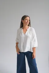 Blusa blanca de corte holgado con cuello redondo fruncido y abertura frontal en V. Presenta mangas tres cuartos con puños elásticos y detalle de tejido texturizado en los mismos.