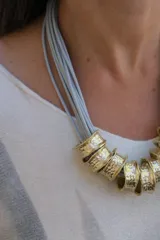 Collar de diseño artesanal compuesto por múltiples tiras de cuero gamuzado en color gris claro, adornado con una serie de piezas cilíndricas de metal dorado con textura martillada.