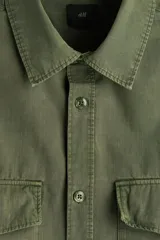 Camisa cargo color verde militar de corte regular, confeccionada en tejido ripstop de algodón. Cuenta con cuello inglés, cierre frontal de botones, bolsillos con solapa en el pecho y mangas largas con puños abotonados.