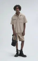 Camisa de manga corta color beige, estilo técnico, confeccionada en microfibra liviana. Cuenta con doble bolsillo frontal, uno con detalle de malla y otro con cierre, y broches en lugar de botones.