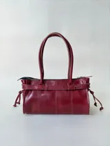Cartera de cuero de tamaño medio con estructura rígida. Presenta una tira decorativa con tachas y hebillas metálicas en los extremos, además de lazos laterales. Cuenta con doble asa para llevar al hombro y cierre superior.
