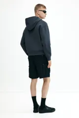 Campera con capucha de algodón pesado, color azul oscuro o gris pizarra, con cierre frontal bidireccional y bolsillos canguro. Presenta un corte holgado (boxy) con hombros caídos y puños y bajo acanalados.