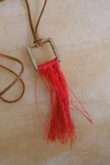 Collar con cordón de cuero tostado y dije geométrico metálico de sección cuadrada con flecos colgantes de hilo rojo.