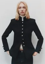 Chaqueta estructurada de corte recto en color negro, con cuello mao y cierre frontal de botones dorados con efecto joya. Presenta manga larga con puños abotonados y forro interior.