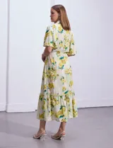Vestido largo camisero con estampado floral amarillo y verde sobre fondo blanco. Tiene cuello camisero, mangas cortas con volados, cintura con lazo ajustable y falda escalonada con volados.