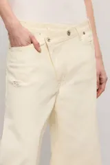 Pantalon de mezclilla color marfil con diseño cruzado asimétrico en la cintura, perneras anchas de corte relajado y detalles de roturas sutiles en la rodilla y el bolsillo.