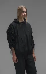 Campera rompevientos negra de calce oversize, confeccionada en microfibra técnica ligera con acabado semi-mate. Presenta capucha integrada, cierre frontal técnico y bolsillos tipo ojal. Destaca por su sistema de frunces elásticos regulables mediante tancas a lo largo de las mangas y el ruedo, permitiendo ajustar el volumen de la prenda.